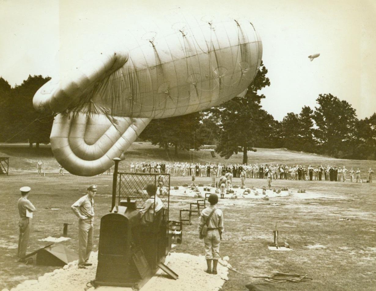 Barrage Balloons Ready In N.Y. Area, 6/29/1943. The busy and populous New York Metropolitan Area is now well protected from low-level enemy air attack, by barrage balloons, which have recently been placed in their battle positions. Under the direction of Maj. Gen. Sanderford Jarman’s antiaircraft artillery command, the balloons offer a menace to any enemy air raiders that try to attack New York City. Completely equipped with a repair shop and gas-generating unit, these “kite” balloons are ready for instant use. When they have been raised, they stretch steel cables that would knife through the wing of any low flying plane like a hot knife through butter. A ring of these “rubber cows” force enemy bombers to fly at high altitudes, lowering bombing efficiency. Furthermore, they are extremely effective against dive-bombing and “hedgehopping” aircraft. In this series of photos, are some of the balloons that now defend New York City. Here, at a balloon station “somewhere on Long Island,” members of the 13-man crew stand by to release a “rubber cow.: in left foreground is the power winch which raises or lowers the balloon by means of its steel cable. Balloon can be raised several thousand feet in a matter of seconds. 6/29/43 (ACME);