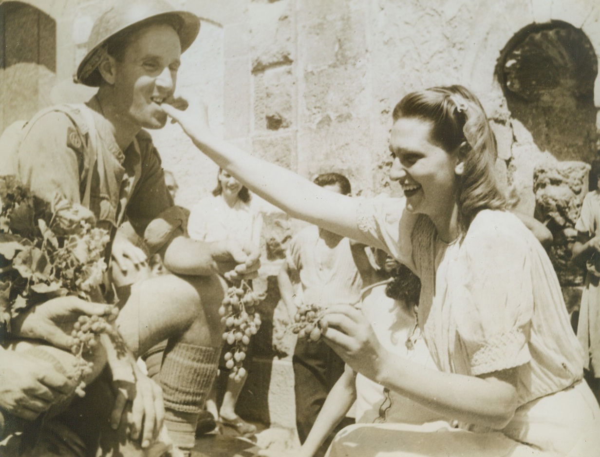 Grapes for the Victors, 8/29/1943. CATANIA, SICILY—This British Tommy grins as a pretty Sicilian girl feeds him grapes and a great big smile to welcome him and his Eighth Army buddies to a village new Catania. Although they were unable to speak English, happy Sicilian women conveyed their greetings to the victors with flowers, fruit and smiles.  Credit: ACME.;