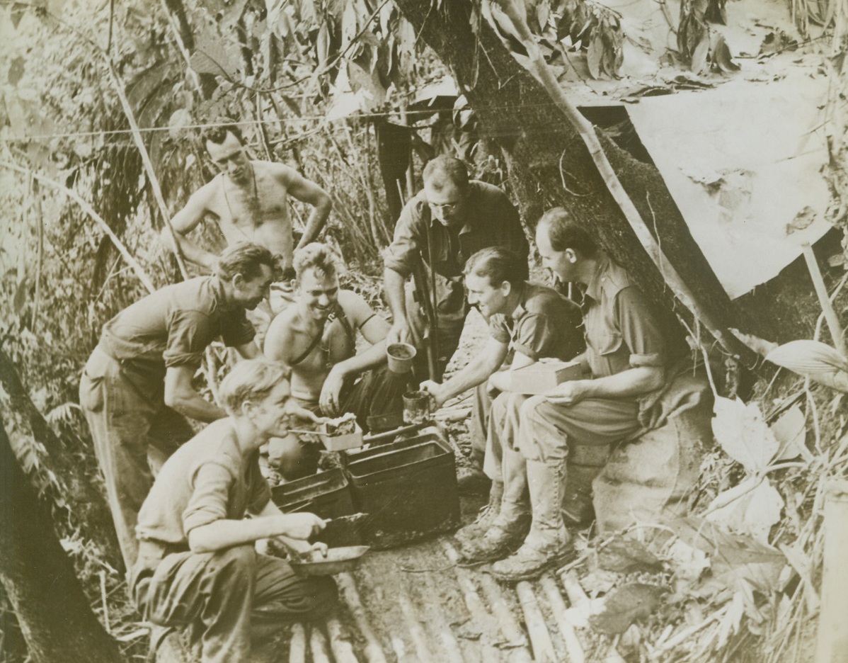 YANKS, AUSSIES CLOSE IN ON SALAMAUA, 9/2/1943. NEW GUINEA—Yanks and Aussies enjoy a hot meal at this out door kitchen on a mountainside near Mubo. The Allied units to which these men belong are closing in on the Jap air field at Salamaua, which is expected to fall at any hour. Left to right, are: Pvt. Les Woolley, of Footscray, Victoria; Pvt. Les Oates, Wangaratta, Victoria, (the cook); Pvt. Bern Buck, Hendigo, Victoria; Sgt. Roy Young, Portland, Oregon; Warrant Officer Sim Woodcock, Fongala, Victoria, Pvt. Frank Bellett, Donald, Victoria; and Staff Sgt. Leo Bidge, of Deniliquin, N.S.  Credit Line (ACME);