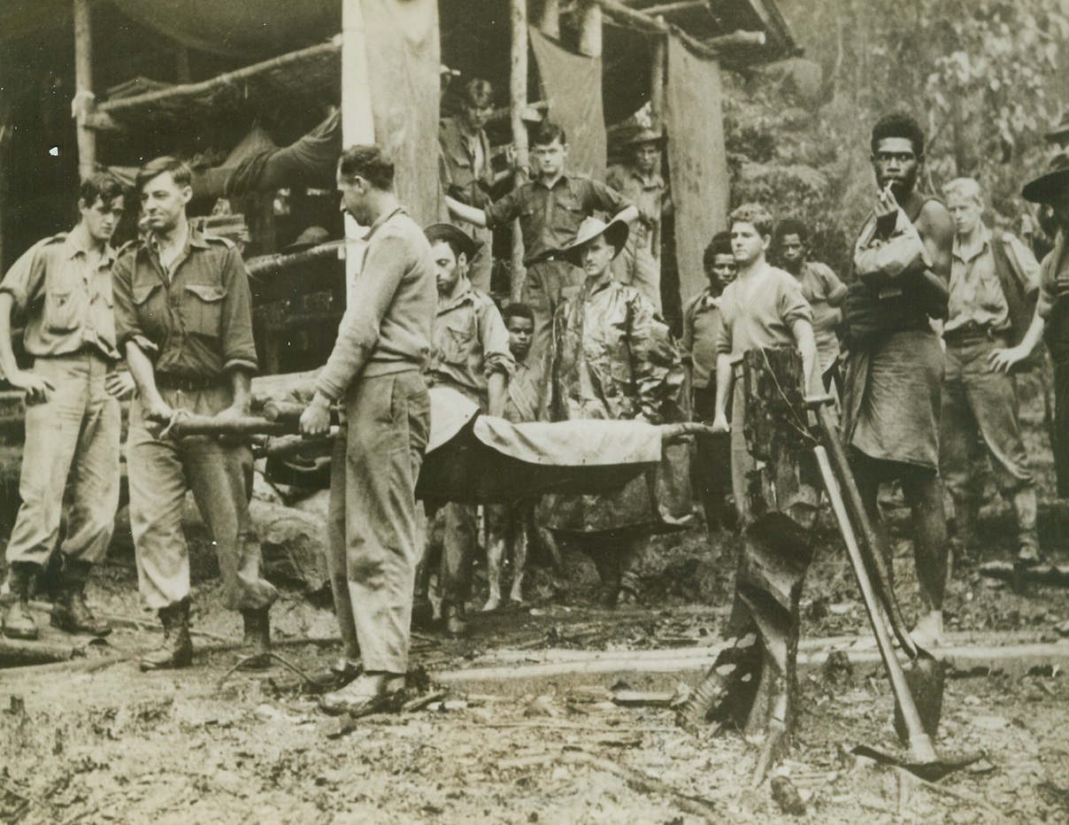 AMBULANCE AFOOT, 9/14/1943. New Guinea—A wounded American soldier is carried from the advance dressing station at House Banana, somewhere the New Guinea, before being turned over to natives who will shoulder the stretcher to the next day’s staging camp, a day’s walking distance away. This camp is on the track several thousands of feet up in the mountains of the Tambu area of New Guinea where there is a mist at some time each day. Here, the mean rest in tiered bunks and the walls are covered with blankets.  Credit: ACME.;