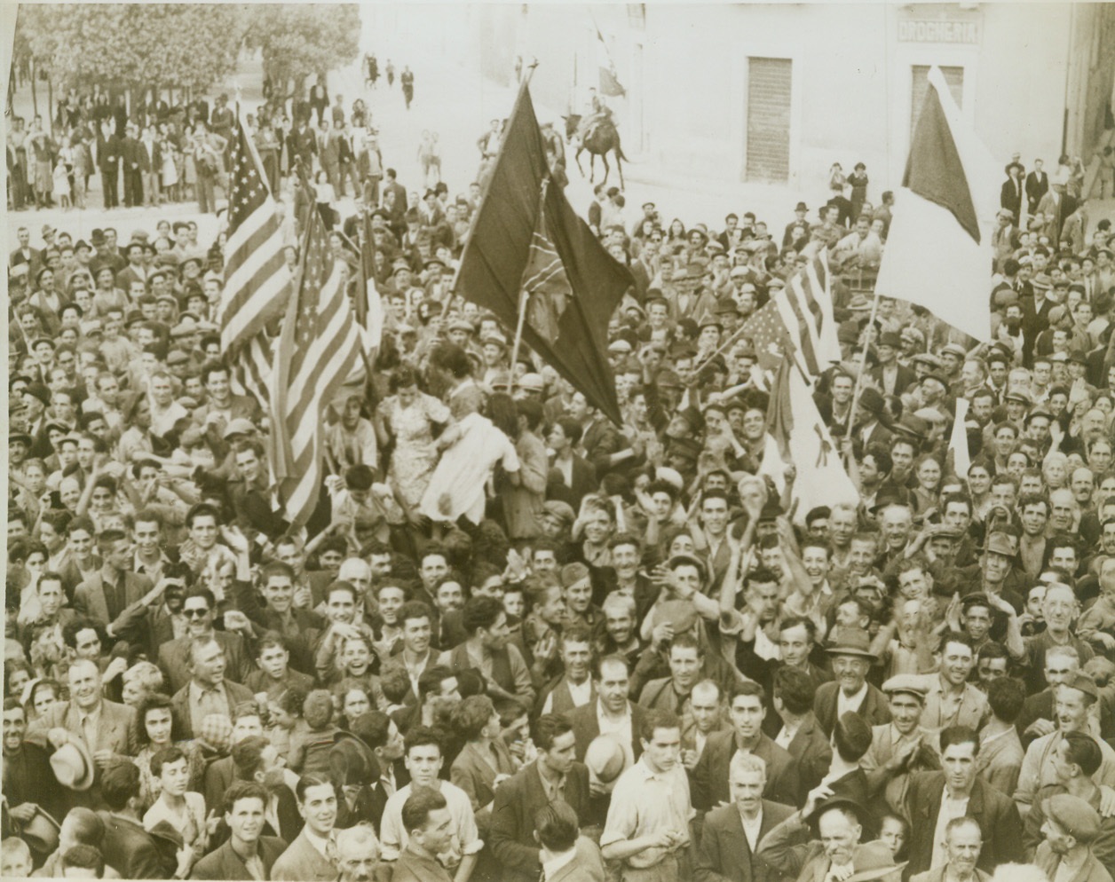Welcome – Italian Style, 10/29/1943. Italy – American, British and Italian flags were held aloft by this crowd of Italians in the village square, welcomed Canadians to Gravina. Men of a Western Canada reconnaissance unit, first into Gravina, received a tremendous welcome. Credit: (ACME);