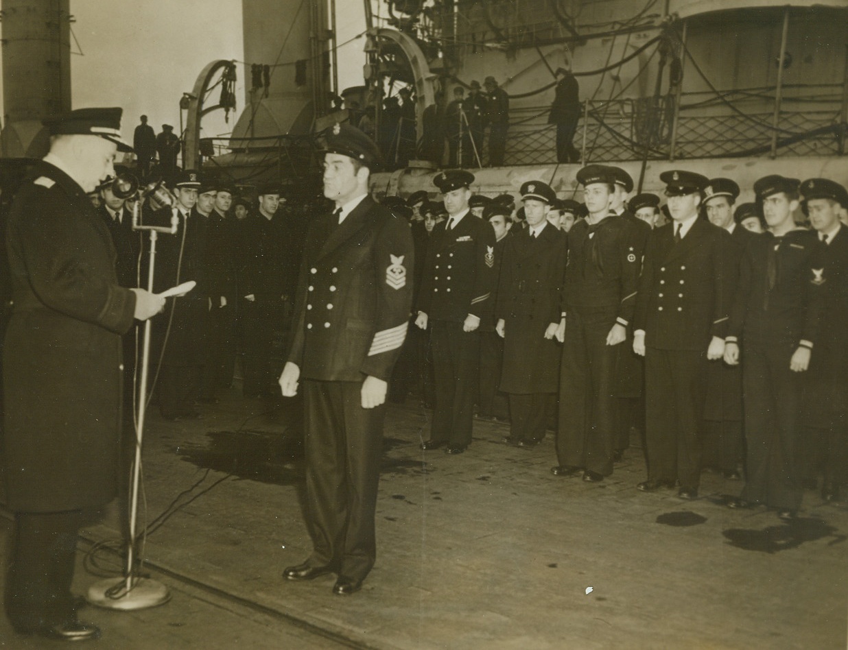 Honored for “Extraordinary Heroism”, 11/22/1943. Chief Water Tender James William Daughtery of Abilene, Texas, stands at attention as the citation awarded him the Navy Cross is read. Daughtery’s ship, a destroyer (seen in background) was struck by an aerial bomb at Palermo last August, and badly damaged. “With complete disregard for his own safety (he) cut away a blackout curtain and entered a fire room and assisted the trapped men there to safety.” Read Admiral Monroe Kelly, USN, presented the medal.Credit: Official US Navy photo from ACME.;