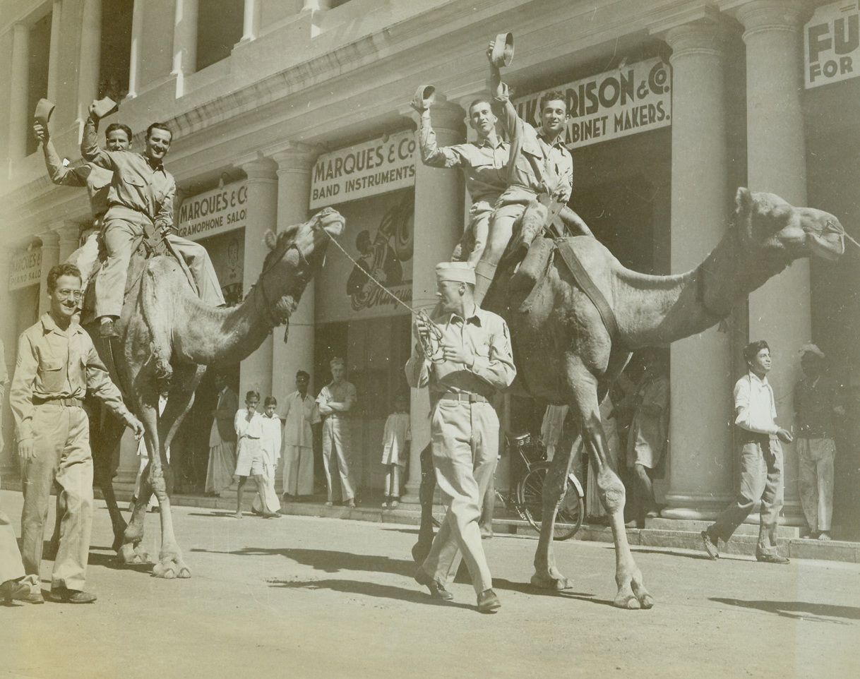 YANKS TACKLE “SHIPS OF THE DESERT”, 12/17/1942. INDIA—American soldiers stationed in India, take time out for a camel ride down the main street of a city, “Somewhere in India,” as natives watch with interest. Credit: ACME.;