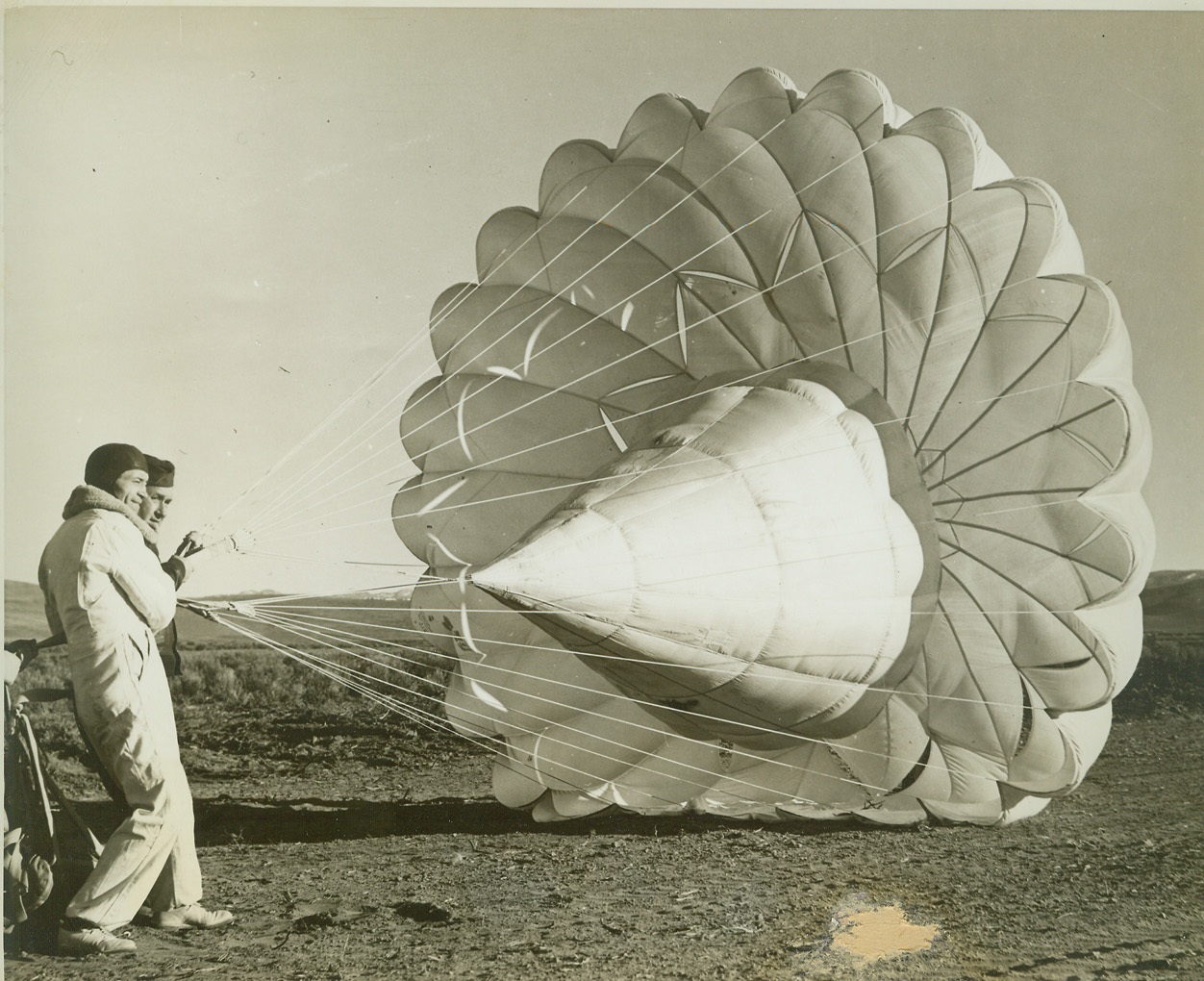 Army Tests New Type Parachute, 3/8/1942. Reno, Nev.—Utilizing a center cone (illegible portion here), opening shock and reduce speed of (illegible portion), a new type parachute is shown after its first Army tests by Jumper George Waltz. In foreground is the inventor, Dr. Christian Wolf. Waltz made the first test jump from 2700 feet. Credit: ACME;