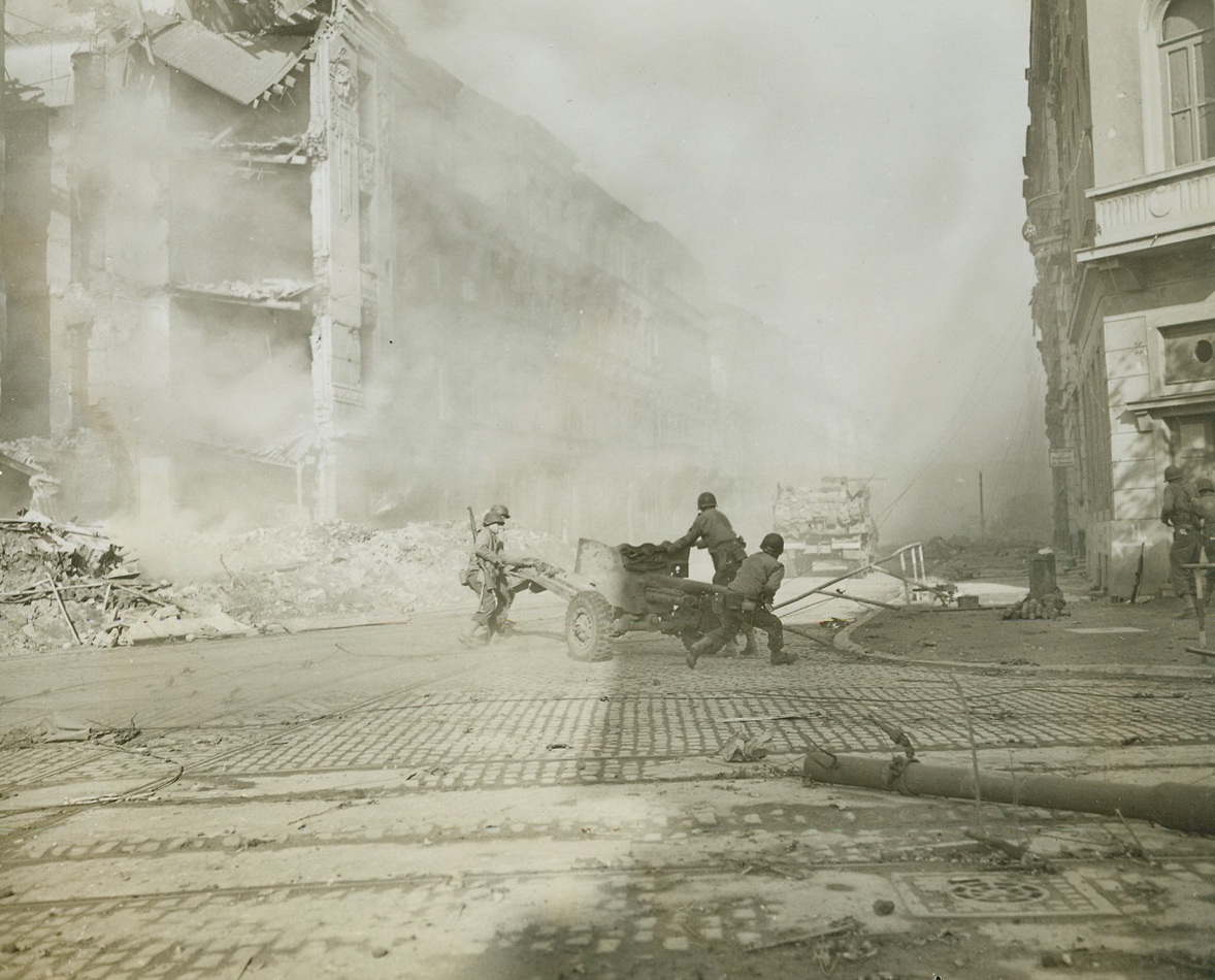 German Key City is Battleground, 10/20/1944. AACHEN, GERMANY -- As the city becomes darkened by a pall of smoke from exploding shell bursts, Yank artillerymen move their gun into position on the corner of Bismarck St. in Aachen, as street-to-street fighting continues in the besieged German city. It was announced today that the American First Army had completed the mop-up of Aachen, leaving Gen. Hodges' Army free to continue its drive eastward over the Cologne plain. Credit (ACME Photo by Bert Brandt, War Pool Correspondent);