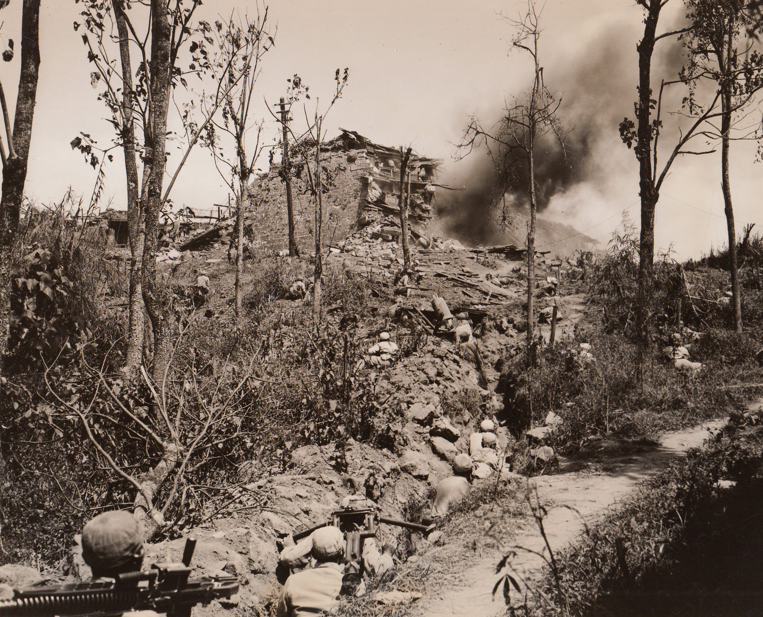 Tengchung Captured from Japs – (#3), 10/4/1944. China – The first city east of Burma to be liberated by the Allies, the ancient jade center of Tengchung has been captured from the Japs after five bloody weeks of fighting. Here, (foreground), Chinese troops wait to move up on the city’s wall, as engineers blast holes through the 20-foot-thick barricade (background). The smoke is from exploding charges.;