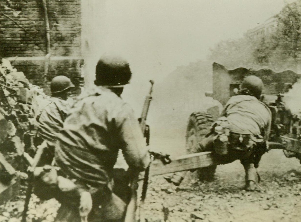 Blasting Away At Aachen, 10/16/1944. American troops direct their light artillery fire at a tower in Aachen, which the Germans used as an observation post. The tower may be seen through the haze directly to the left of the muzzle of the infantryman’s rifle in the center of the picture. (Radiotelephoto);