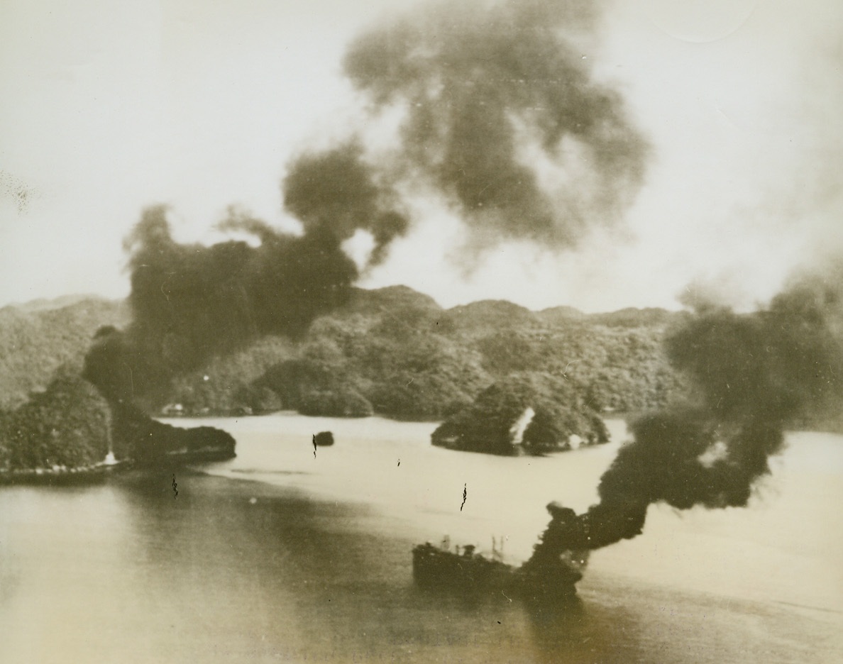 No Title. 4/15/1944. Palau Islands – Twin columns of thick, black oil smoke rise from a pair of burning Jap ships which were sunk off the shores of Palau by carrier planes of a U.S. Navy Task Force raiding the important enemy base on March 29th and 30th.  The burning ship in foreground is a large cargo vessel.  The other target is hidden by a tiny island at left.Credit Line (Official U.S. Navy photo from ACME;