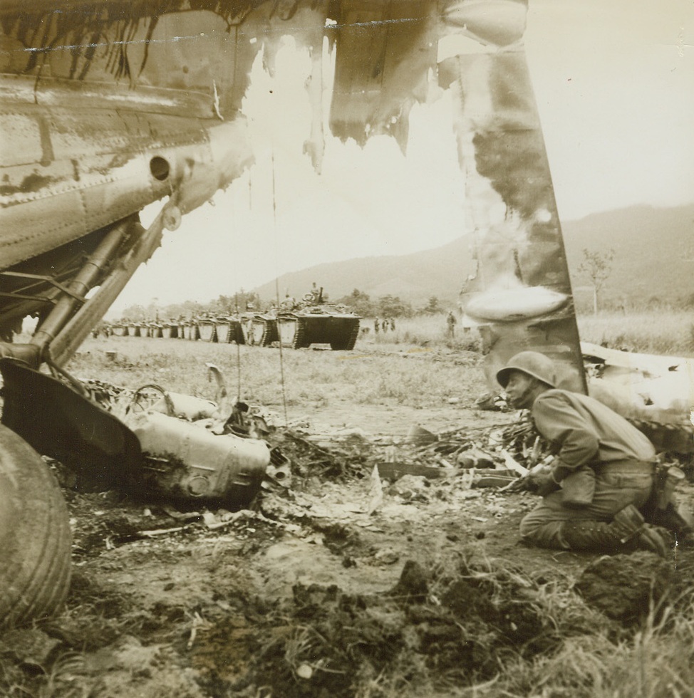 Alligators thru the Wreckage, 5/18/1944. Hollandia, Dutch New Guinea – In a line stretching far as the eye can see, amphibious Alligator Tanks are shown through the wreckage of a Japanese plane on Sentani Airdrome, near Hollandia. The alligators helped to take the airfield by circling through Lake Sentani. The soldier in foreground is apparently on the lookout for any Japs left in the area.Credit: ACME photo by Frank Prist, Jr. for the War Picture Pool;