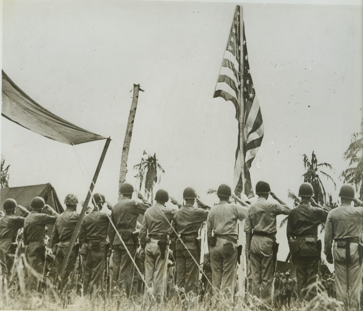 The Call to Colors, 8/6/1944. Guam – Still attired in their battle helmets, Marines of the First Provisional Marine Brigade snap to a smart salute as the American flag waves aloft in the first flag-raising ceremony in front of Maj. Gen. Roy S. Geiger’s headquarters at Agat, Orote Peninsula. Credit (ACME);