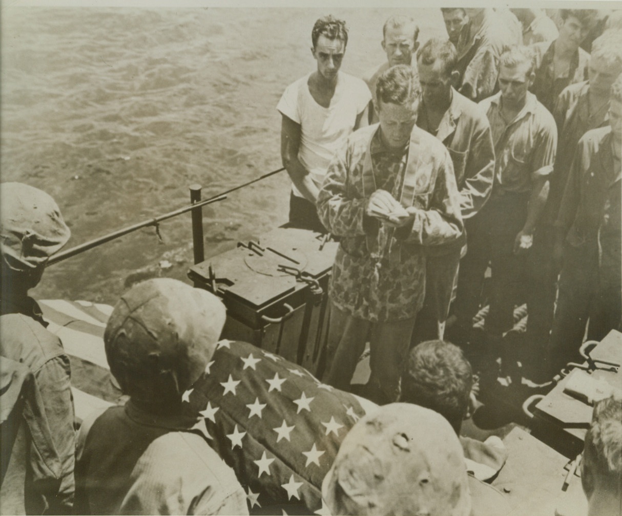 LAST RITES FOR MARINE, 8/8/1944. SOMEWHERE IN THE PACIFIC – Still wearing his camouflaged tunic, Lt. (JG) Joseph P. F. Gallagher, USNR, Catholic Chaplain, 840 Grand Concourse, Bronx, N.Y., reads the burial service for a Marine who died in the assault on Tinian Island. His friends form an Honor Guard for the burial at sea. Credit: U.S. Marine Corp photo via OWI Radiophoto from ACME;