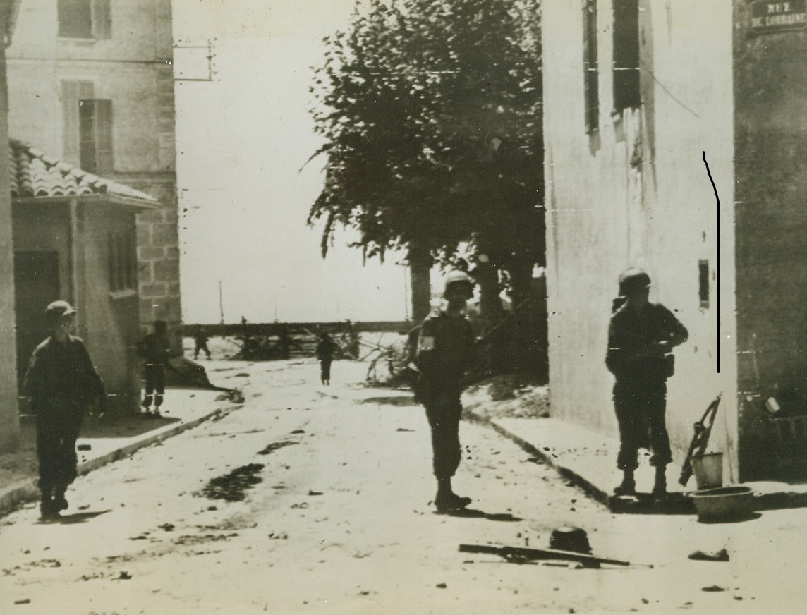 Expanding New Beachhead, 8/16/1944. Moving cautiously and watching every house for enemy snipers, American Infantrymen advance through a street in a town east of Toulon. In the background, can be seen the beach, one of the many points Allied invasion forces hit along the coast of Southern France. Note German rifle and helmet (right, foreground). Credit: ACME PHOTO BY SHERMAN MONTROSE FOR WAR PICTURE POOL VIA ARMY RADIOTELEPHOTO.;