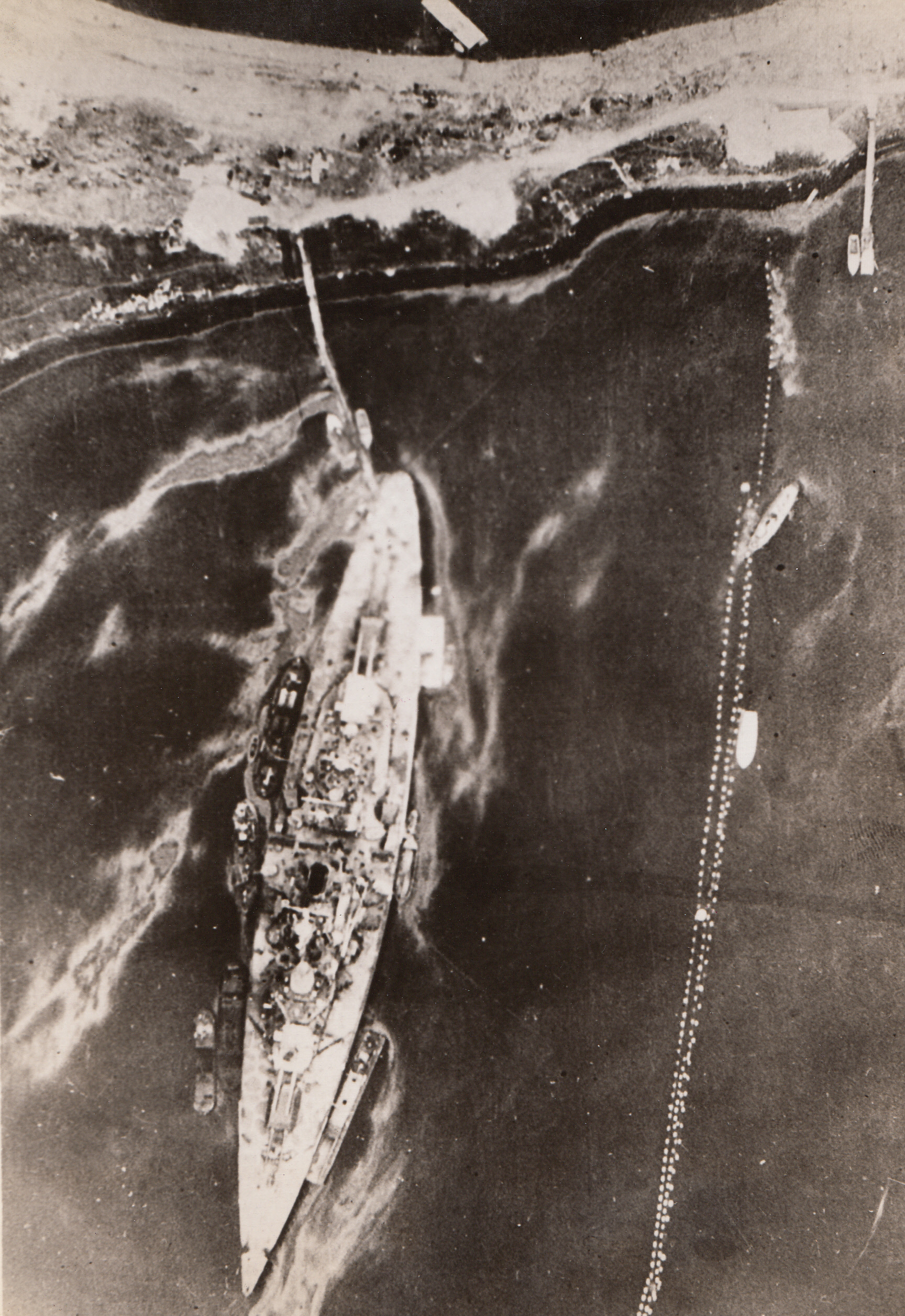 Twice-Damaged Tirpitz, 8/12/1944. NORWAY – This recent photo of the Tirpitz at her moorings in Kaafjord, Norway, was taken by an RAF reconnaissance plane of the Coastal Command. The ship was damaged in Kaafjord on two occasions, once by British submarines on Sept. 22, 1944, and again by Fleet Air Arm attack on April 3, 1944. Some of the damage has evidently been repaired, but the starboard boat and aircraft crane are missing.;