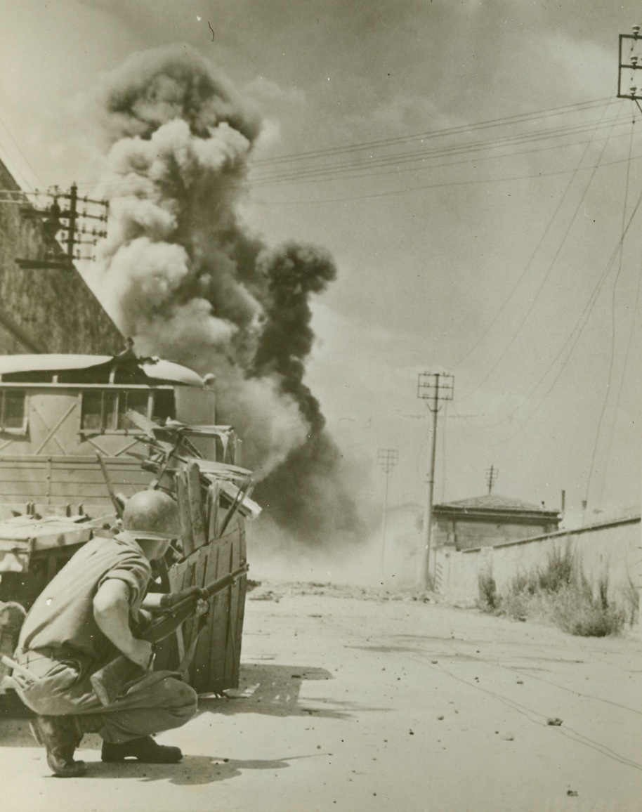 Enemy Mines Go Up, 8/7/1944. LEGHORN, ITALY – Taking cover behind a tank, Cpl. Duane T. Moen, Minneapolis, Minn., is on the alert for snipers as his Fifth Army buddies set off mines sowed heavily in one of the main streets leading to Leghorn harbor. A giant column of smoke rises in background as the mines blow up.Credit Line (Official U.S. Army Photo from Acme);