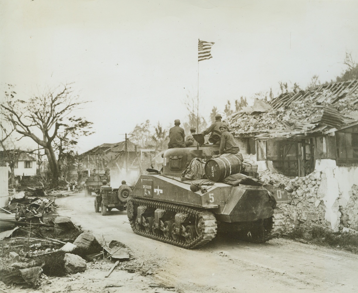 Old Glory Moves Up to Battle, 8/11/1944. GANA, GUAM – Moving through the war-shattered town of Agana, tanks and armored vehicles of the Third  U.S. Marine Division roll to the North on the hells of retreating Japs. They carry with them the American flag, to wave once again over the island that was seized by the Japs shortly after Pearl Harbor. Credit: ACME;