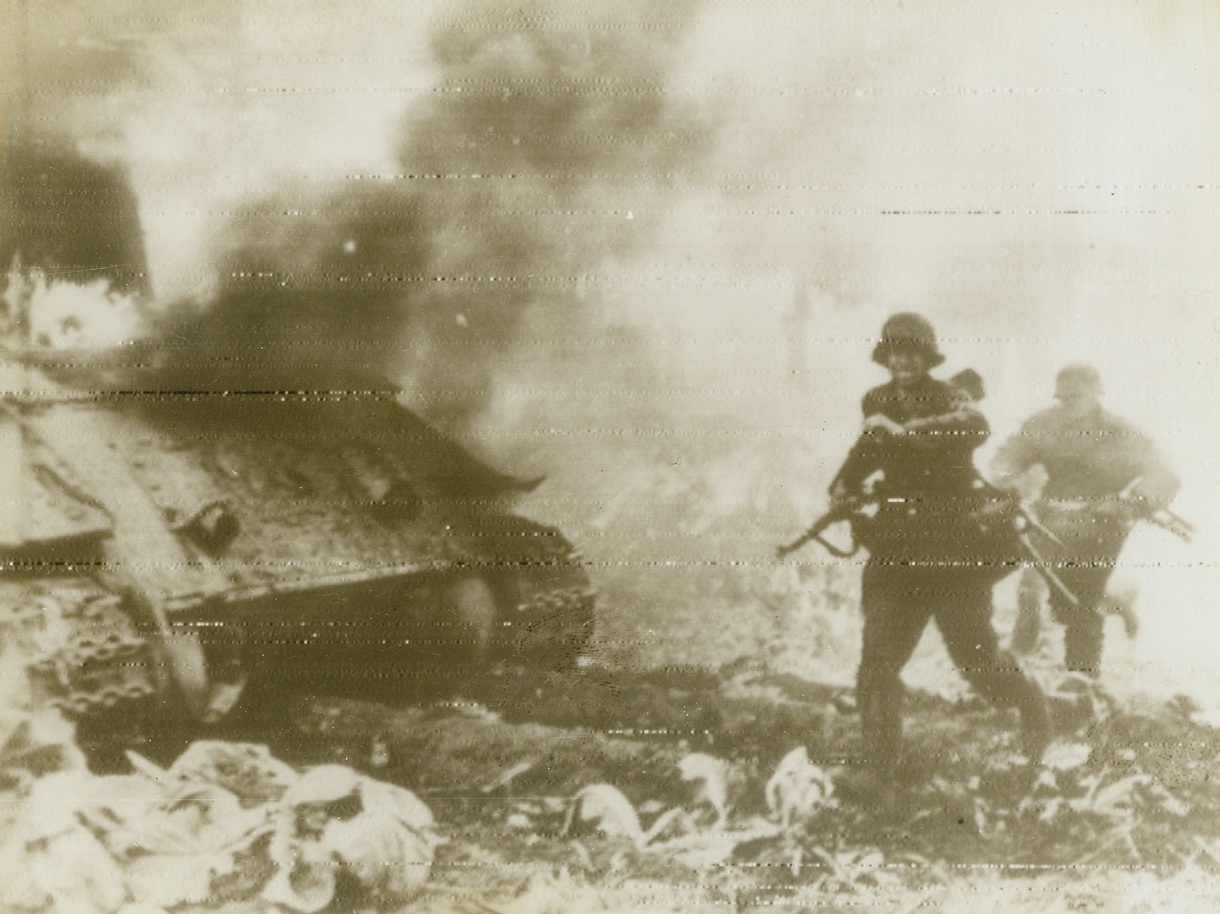 In the Midst of Battle, 8/23/1944. On the Russian Front—A German Panzer Grenadier dashes past a flaming tank during bitter fighting on the Eastern front. The tank was blasted by Russian trench mortar fire. Photo radioed to New York today (8/23) from Stockholm.  Credit: ACME radiophoto.;