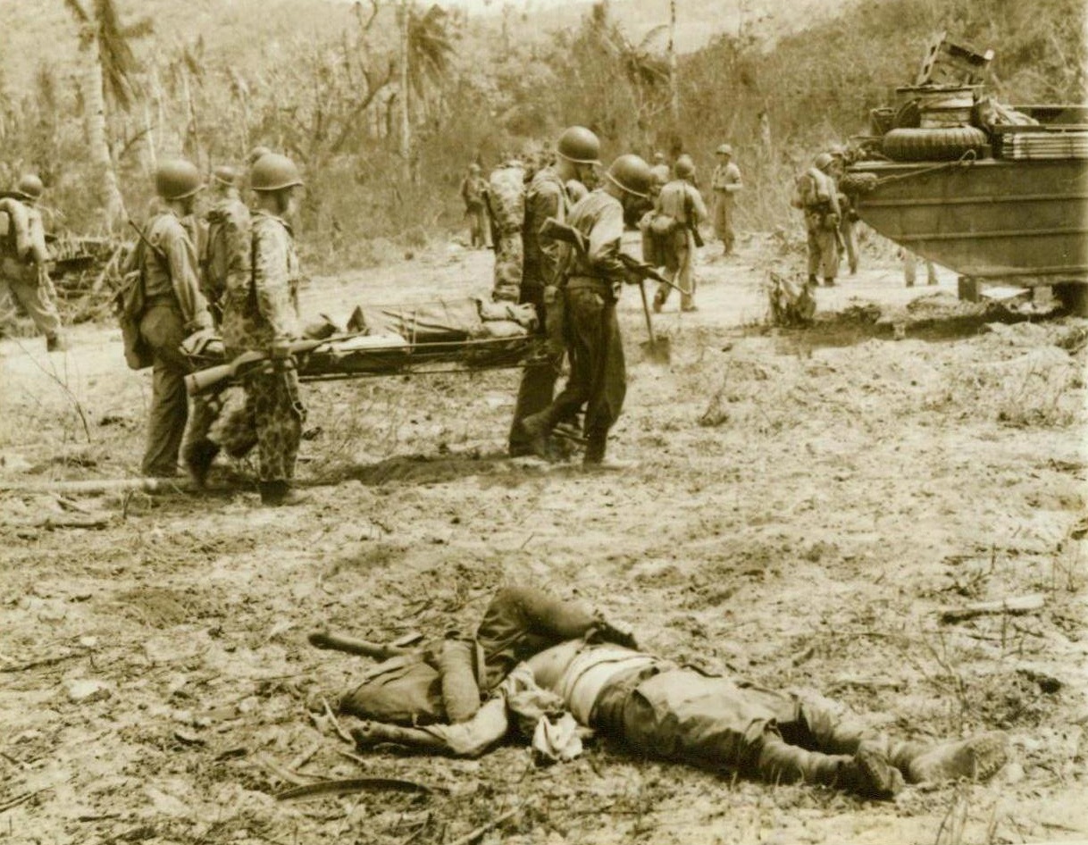 One of Many, 8/4/1944. Guam—US Marines move forward near Asan Point, carrying a litter of supplies to their forward troops. In the foreground is a dead Jap, one of the 7,419 enemy dead who have been buried by the Marines since they landed on the island. Although enemy resistance has stiffened, the American forces continue to advance unchecked. 8/4/44 (ACME);