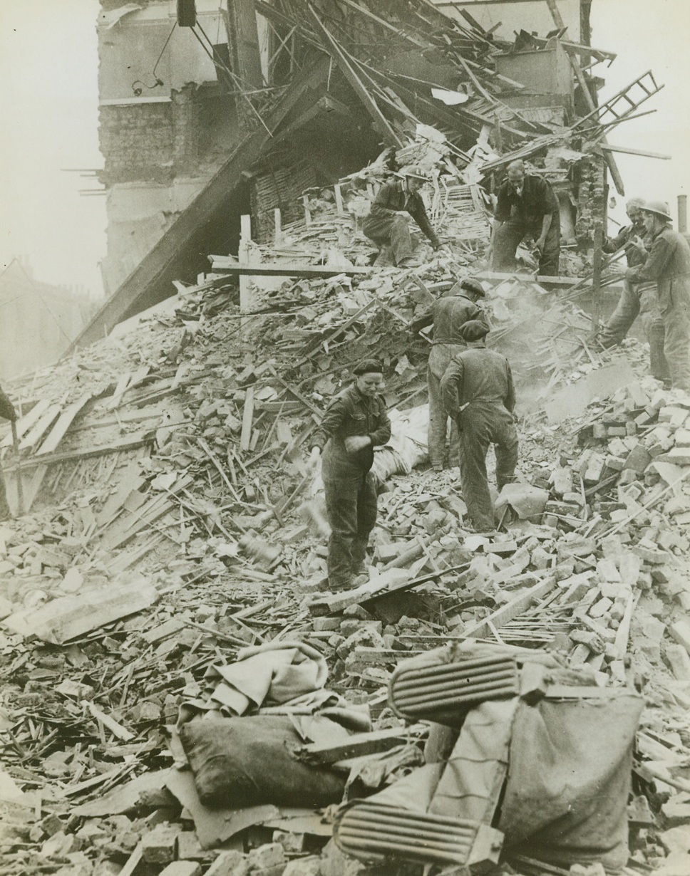Robot Bombs Devastate Southern England, 8/7/1944. Southern England – While falling concrete and plaster raise a smoky haze, workmen search through the wreckage of homes for victims of the raid. A direct hit from one of the bombs has completely razed this house and only a wall of the one in the background remains standing. Credit: ACME;