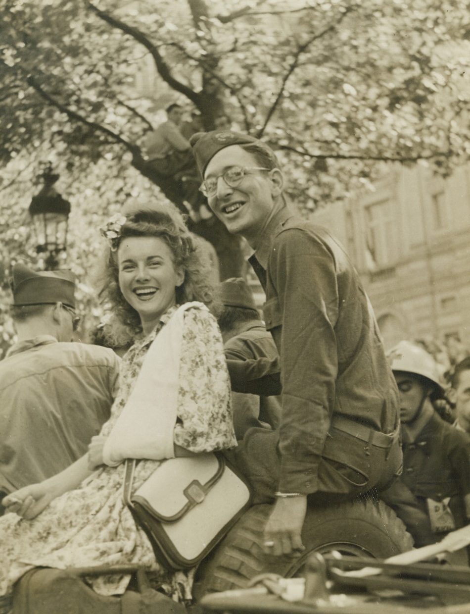 NEA Correspondent Does Himself Proud, 8/30/1944. France – Tom Wolf, NEA war correspondent, sits with his arm around a very beautiful French lassie whose heart he probably captured by offering her a lift in his jeep as liberating forces entered Paris. The blonde-haired beauty has her arm in a sling. Credit: ACME Photo by Bert Brandt, War Pool Correspondent;