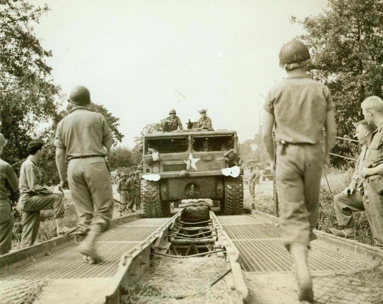 Rebuilding As They Go, 8/21/1944. France – On the heels of rapidly advancing American forces in France, Yank engineers are quickly repairing and rebuilding roads and bridges wrecked by the retreating enemy. This prime mover pulls artillery equipment over a treadway bridge speedily erected by the Americans. 8/21/44 (ACME Photo By Bert Brandt  For The War Picture Pool);