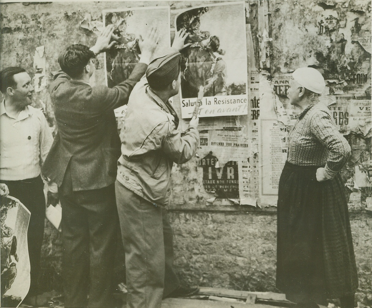 Down With the Old Up With the Free, 8/22/1944. FRANCE -- Residents of Columbierre help an American soldier cover the remnants of Nazi display propaganda with Allied posters. An elderly French woman surveys the work with satisfaction. Credit (OWI Photo from ACME);