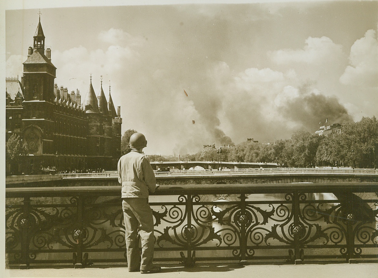 Across the Bridge Paris Burns, 8/30/1944. France—An Allied soldier stands on one of the many bridges across the Seine in Paris to watch fires burning on the outskirts of the French capital during the spasmodic fighting that took place before the city was completely liberated.  Credit: ACME photo by Bert Brandt, War Pool correspondent;