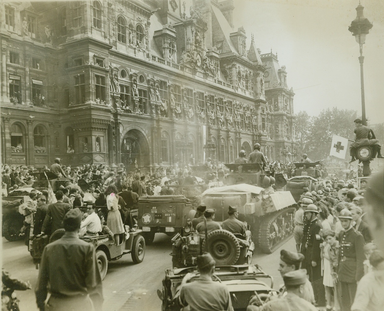 Stage Set For Panic, 8/28/1944. Paris—This was the scene in the Place de L’Hotel de Ville before snipers sent a panic-stricken crowd scurrying for their lives during the official reception for General Charles de Gaulle. The street is jammed with Allied armored vehicles and hundreds of celebrating Parisians. At left, spectators jam the windows of the Hotel de Ville, while at right one spectator has climbed to a perch halfway up a lamp post to witness the celebration.  Credit: ACME photo by Andy Lopez for the War Picture Pool;