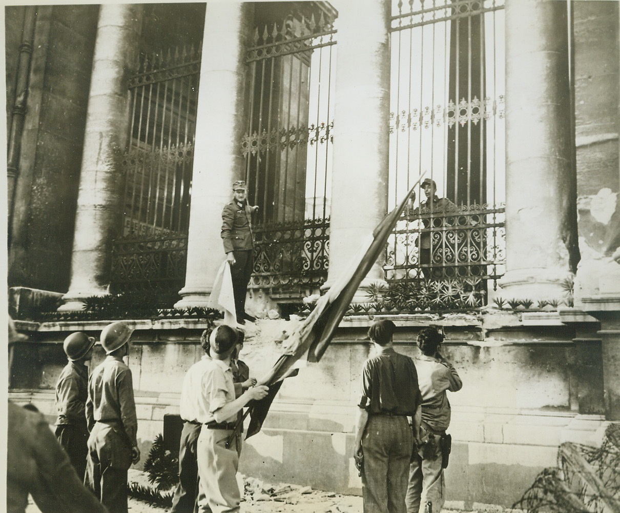 France Regains Its Chamber of Deputies, 8/30/1944. France—With a Nazi garrison barricaded behind him, a German officer hangs onto rail of the Chamber of Deputies in Paris while negotiating for surrender to the French partisans. He holds the white flag of truce while a French soldier in the foreground has the Tricolor ready to hoist as a signal of victory when surrender is complete.  Credit: ACME;