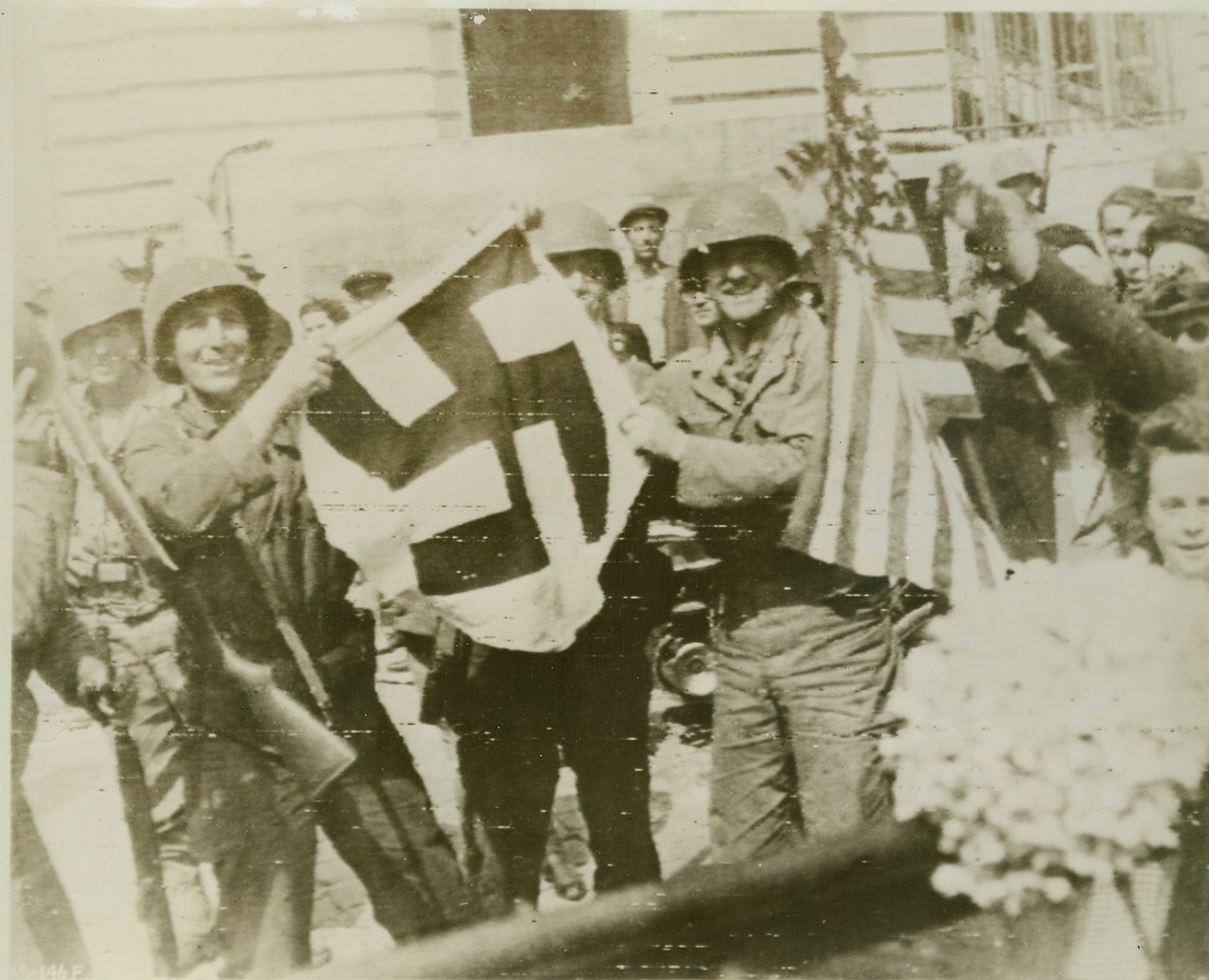 She’ll Wave No More, 8/6/1944. Rennes, France—Smiling Yanks hold the flag that will fly no longer over liberated Rennes, capital of Brittany and largest French town to fall to the Allies to date. Beside the deposed Swastika, someone waves the Stars and Stripes, which, with the Tricolor, now waves over the city. Credit: Signal Corps radiotelephoto from ACME;