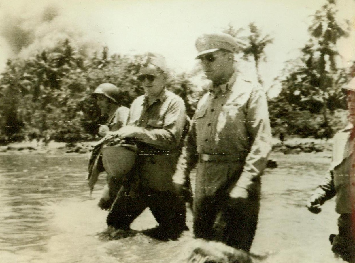 The General on the Scene, 9/19/1944. Morotai, Halmahera Is.—Wading through water knee-deep, Gen. Douglas MacArthur (right) heads for a landing barge as he returns to his ship after inspecting American troops on Morotai. The man on the left is an American Naval Doctor. In the background (Left) Smoke rises from a burning Jap oil dump. 9/14/44 (Acme);