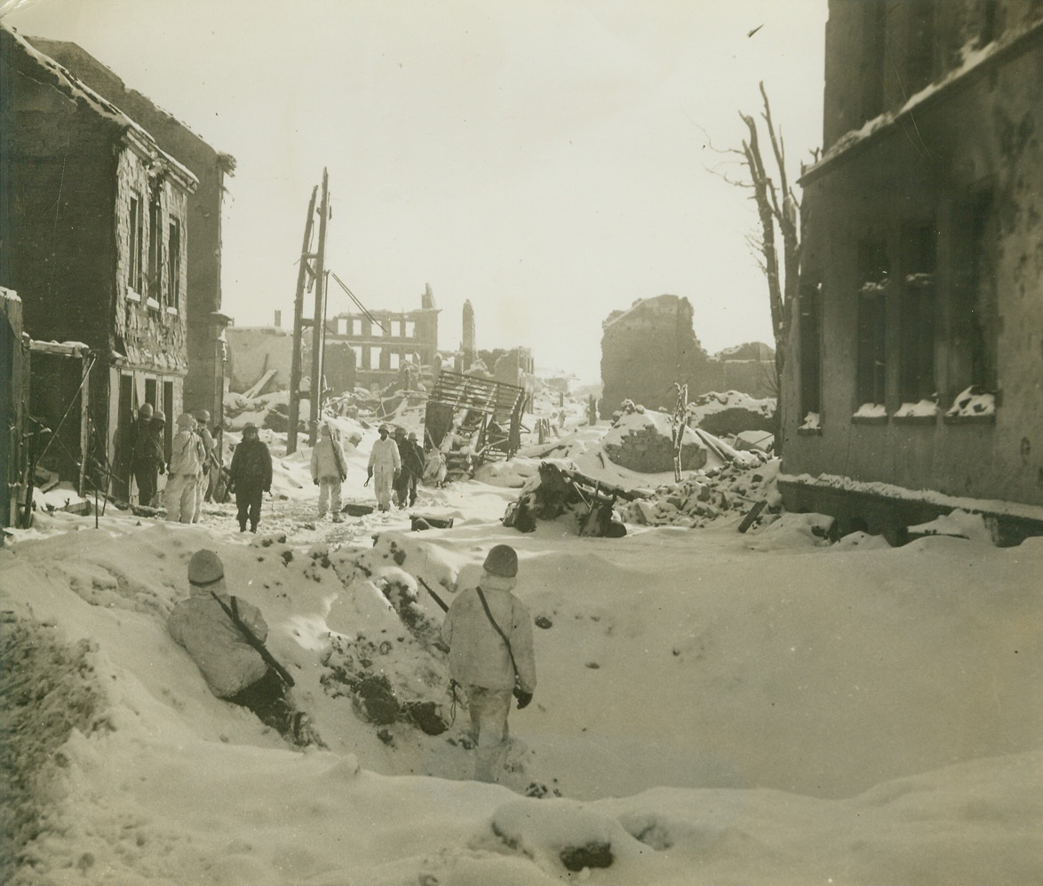St. Vith Street Scene, 1/30/1945. BELGIUM – Two U.S. First Army soldiers, in snow-covered crater, cover their comrades patrolling the streets of St.Vith, Belgium, for possible enemy snipers. The city, a vital road junction in the now eliminated Belgian Bulge, was found completely ruined when re-captured from the Nazis. Skeleton homes can be seen at left and in background. Photo by Acme photographer, Harold Siegman, for the War Picture Pool. Credit – WP – (Acme);