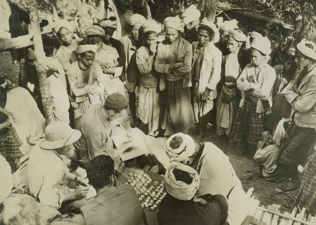 NAGA PAY DAY, 2/10/1943. SOMEWHERE IN INDIA—Nagas eagerly crowd around the stacks of silver rupees in which they are paid for their work assisting the U.S. troops in India. The Naga chief and his assistant sign regular U.S. Army pay vouchers by inked thumb prints. These natives, who move supplies, refuse paper money. Watching the dark-skinned workers get their coin are Pfc. Richard Maccio, East Rochester, N.Y. (foreground, back turned); Sgt. Milton F. Elkins, Haskell, Texas (sun helmet left) and Sgt. Wm. L. Dean, San Francisco (sun helmet), who works on the pay vouchers. Credit: Official U.S. Army photo from Acme;
