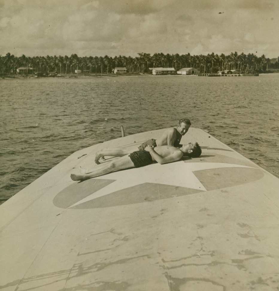HO-HUM, 2/12/1943. SOUTH PACIFIC—Two American fighters, based aboard “The Seaplane Tender,” stretch out on the wing of a plane and lap up a little South Pacific sun. Their plane is one of many on the giant floating repair station which has shops and equipment for the complete overhauling of aircraft. Pilots and crews live aboard the sea-based garage and do might well, too, for the ship has a barber shop, canteen, soda fountain and even a laundry. Credit Line (ACME);