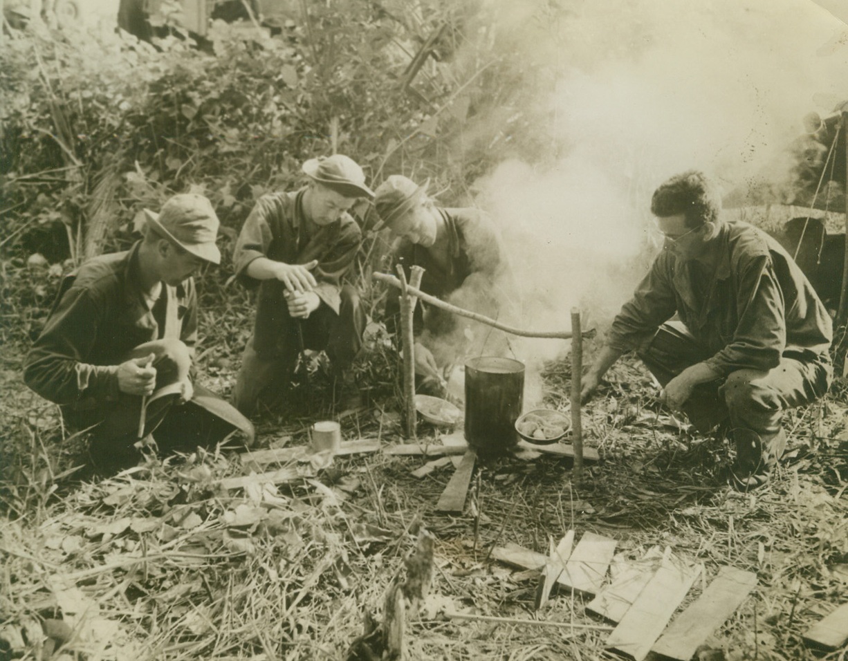 CHOW FOR YANKS IN BUNA, 2/10/1943. BUNA, NEW GUINEA—Soldiers of the 32nd United States Division, take time out from fighting Japs in the Buna sector, to attack their field rations, which they heat over a small fire. Left to Right, are: Pvt. Joseph F. Bitner, of Garwood, Texas; Pfc. Richard J. Leddy, of Detroit, Mich.; Pvt. Lewis M. Hawes, Aberdeen, Idaho, (who was later reported missing) and Pvt. Rodney F. Corter, Tucson, Ariz. Since this photo, which was released in Washington today, was taken, American and Australian forces have cleaned the Japs out of Buna, killing some 15,000 of them. Yesterday, Tokyo finally admitted that the Japanese had “withdrawn” from Buna. Credit Line (U.S. Army Photo from ACME);