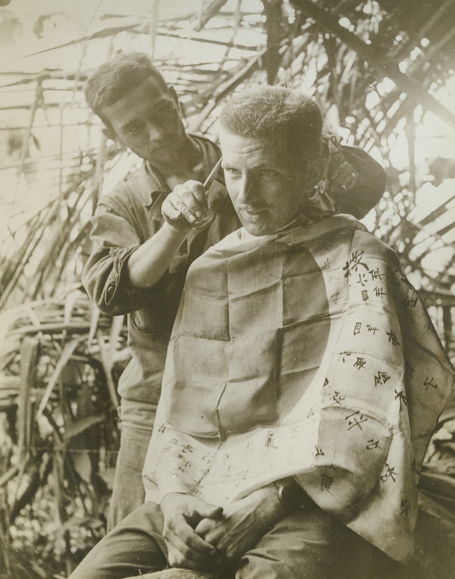 LET THE HAIRS FALL WHERE THEY MAY, 2/6/1943. NEW GUINEA—This soldier by isn’t proud. He’ll use any old rag for a barber’s apron while he gets his hair cut—and so he drapes a Jap flag about his shoulders to catch the falling hairs. Photo was made during the Buna campaign. Recent reports from New Guinea indicate that Allied forces have routed the Japs from Papua and are now pounding Jap positions at Lae. Credit: ACME.;