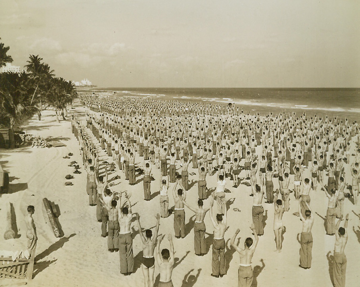 Calisthenics on the Beach, 2/5/1943. Miami Beach, Fla.—Soldiers training with the Army Air Forces at Miami Beach carry out their daily program of calisthenics on the beach. Hundreds of men, exercising along long stretches of the beach, do the same exercises at the same time. Their routine is directed by one announcer whose voice comes to them through loudspeakers erected on the beach.Passed by censors.Credit: ACME.;