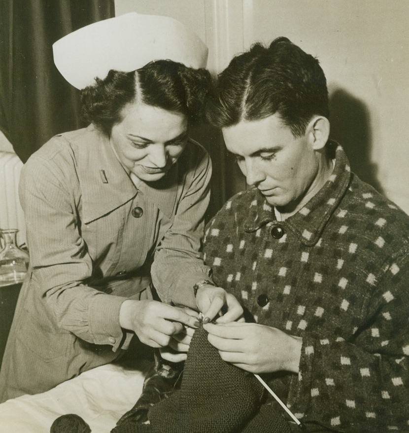 Turnabout, 3/6/1943. ENGLAND – The womenfolks back home in Kansas, have probably been knitting sweaters and socks for him since he went into the Army, but now Pfc. Huey H. Stidham is taking a hand at the needles. 2nd Lt. Gladys Swayze, of Long Island City, N.Y., is giving him the lowdown on the knit one’s, purl two’s in the occupational therapy ward of an American Army Hospital somewhere in England. (Passed by Army Censor) Credit: ACME;