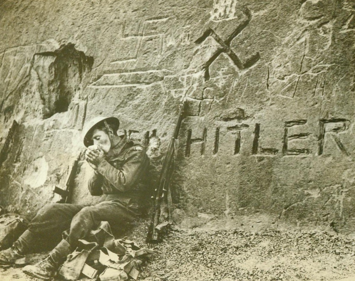 A Bit of Chiselling for Der Fueher, 3/2/1943. WESTERN DESERT - Some exponents of Nazi art, with more enthusiasm than ability, had time to decorate this rock wall with Hitlerian homelies before the British 8th Army chased the Germans out of Libya. None of the artists, however, seemed to know the correct way to reproduce the swastika - they're all wrong. A British Tommy nonchalantly lights a "fag" as he leans against the wall, which is "on the way to Tripoli.";