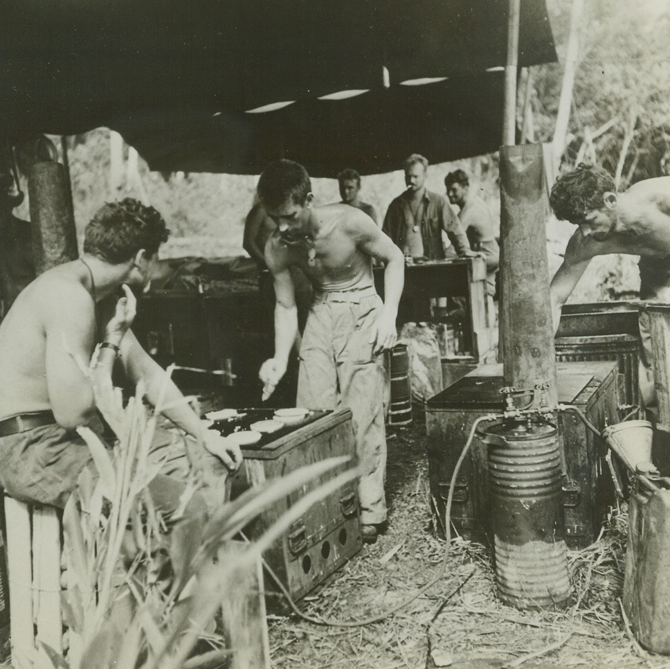 FLAPJACKS FOR A MAN-SIZED APPETITE, 3/7/1943. GUADALCANAL—During the fierce fighting months when the Americans were showing the Japs our new “get them out of here in a hurry” maneuver, the boys on the front lines didn’t have the time to stop and cook. However, they had the situation well in hand as shown in this photo of American cooks making the old American standby—flapjacks, to be sent in hot containers to the front line troops.  Credit: ACME;