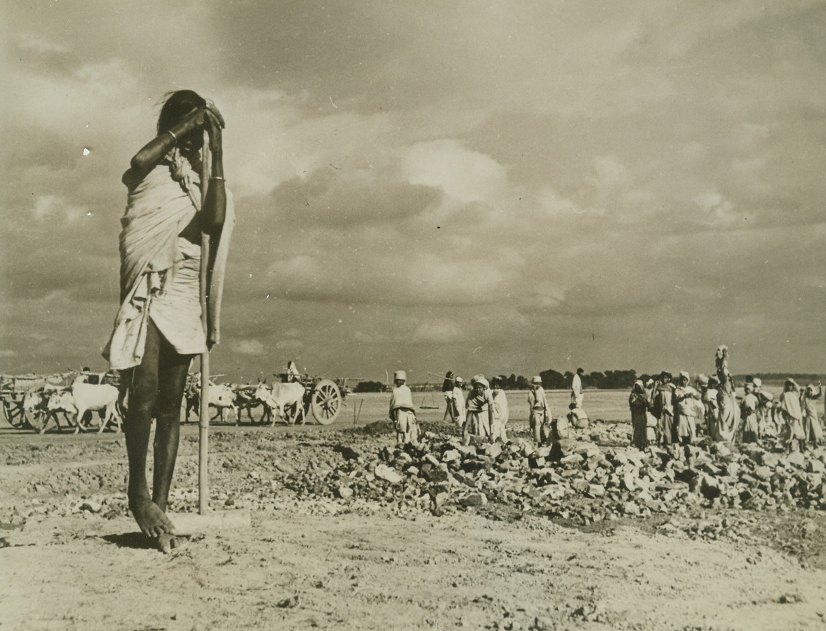 PRIMITIVE LABOR CREATES MODERN WAR TOOL, 3/9/1943.  EASTERN INDIA—A young Indian girl rests on her crude work tool, as other native women balance broken stone on their heads and bullock carts and hand labor carry out their painstaking parts in creating a modern airfield in Eastern India for U.S. planes. The carting and leveling of broken stone is a picture out of an ancient world. Soon, modern fighter, bomber and transport planes will utilize the landing strip. Credit: ACME.;