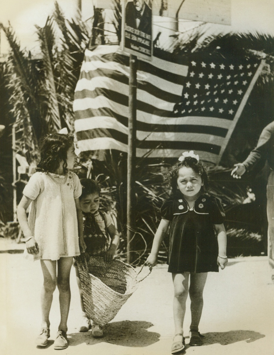 Bringing Home American “Bacon”, 5/21/1943. Tunisia—Three Tunisian youngsters anticipate full tummies as they carry off American foodstuffs distributed at the football stadium in Sousse, Tunisia. A good quantity of the enormous tonnage of U.S. supplies for civilians which has been shipped to North Africa recently has already been distributed to Tunisians, made hungry by the Axis. The shipments consist of goods unobtainable in Tunisia, such as coffee, tea, sugar, biscuits, clothing, and material for making children’s clothes. The people receive ration cards, to be stamped at the time of purchase.Credit: ACME.;