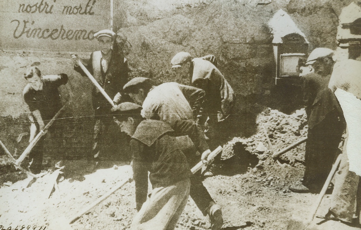 Wanted: More Clean-Up Squads in Sicily, 6/29/1943. SICILY—So great has been the damage done to Sicily by Allied bombers that workmen have been sent over from Italy to aid the clearing of the debris. The poster in back of this street cleaning squad reads: “In the name of our dead we shall conquer.” Sicilians aren’t so sure, a Mussolini orders all but essential workers to evacuate the island’s cities. Photo received in London from a neutral source. Credit: ACME.;