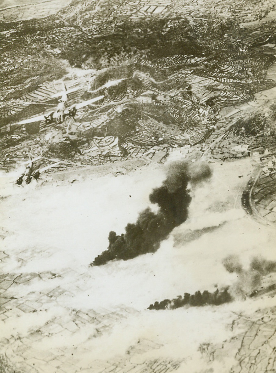 ALLIED BOMBERS SOFTEN UP PANTELLARIA, 6/3/1943. With the fighting in North Africa over, the Allies turn their air force on the Axis ports, airfields and strongholds on the Italian side of the Mediterranean, softening up these strong points in lieu of a possible Allied invasion in that direction. In this photo Baltimore bombers of the South African Air Force drop bombs on the airfield of Pantellaria, Mussolini’s island stronghold between Malta and the African coast, with columns of smoke rising from two oil fires. The landing ground is almost obscured by the smoke of bursting bombs. At the far end of the airfield can be seen the entrances to underground hangars and workshops on the side of the hill. Credit: Acme;