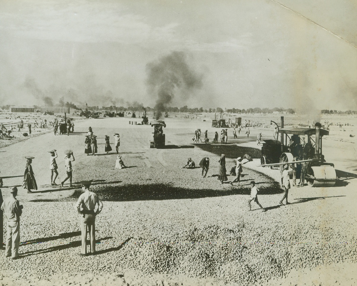 An Airfield Springs Up in India, 6/7/1943. India – Work procedes apace on a U.S. Air Depot somewhere in India.  In surfacing of taxi ramp down past the hangars, a coat of tar is being laid down onto the heavy metalling of rock that lies on top of a heavier foundation of rocks.  The tar is then covered with fine-crushed stone carried on the heads of coolie laborers and cast out over the wet tar.  U.S. officers in the foreground are checking the plans and inspecting the work.  Runway construction and grading of the airfield is done by the Indian Army Garrison Lease Agreement.  Steel hangars in background were brought from the U.S. on the convoy that carried the Task Unit.Credit Line – WP –(ACME);