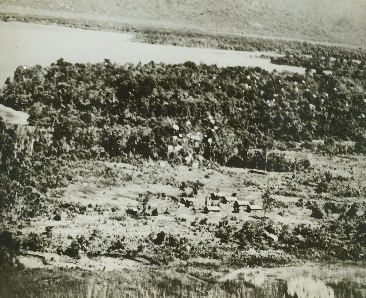 DESTRUCTION VIA PARACHUTE, 6/18/1943. NEW GUINEA—Dozens of heavy bombs float lazily of Japanese installations at Lae, New Guinea, where they were dropped via parachute by U.S. bombers. White spots in the middle right of the picture, and in upper left, show still more bombs which will explode a few feet above the ground, or upon contact, with terrific and devastating effect. This new method of sending bombs via parachute is one of the latest developments in air fighting. Its anti-personnel results are telling, since the downward force of the blast penetrates huts and foxholes and seeks out hidden enemy installations. Credit: U.S. ARMY AIR FORCE PHOTO FROM ACME.;