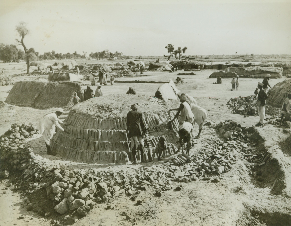 They Help Build a U.S. Airfield, 6/7/1943. India – Here is one of the villages that has been settled near a U.S. air depot for the duration of its construction by Indian workers.   They are tile makers who squatted down in a corner near the end of a taxi ramp.  They did their own clay, draw their own water and with dried grass and cow dung cakes mold and fire the roof tiles for all the barracks and outbuildings that are being constructed under a reverse lend-lease arrangement.  The tiles are fired in the round piles.  The flat floor in the village center is where the tiles are formed out of raw clay and dried in the sun.  Workers and their families live in the grass huts on the edge of the village. Credit line – WP – (ACME);