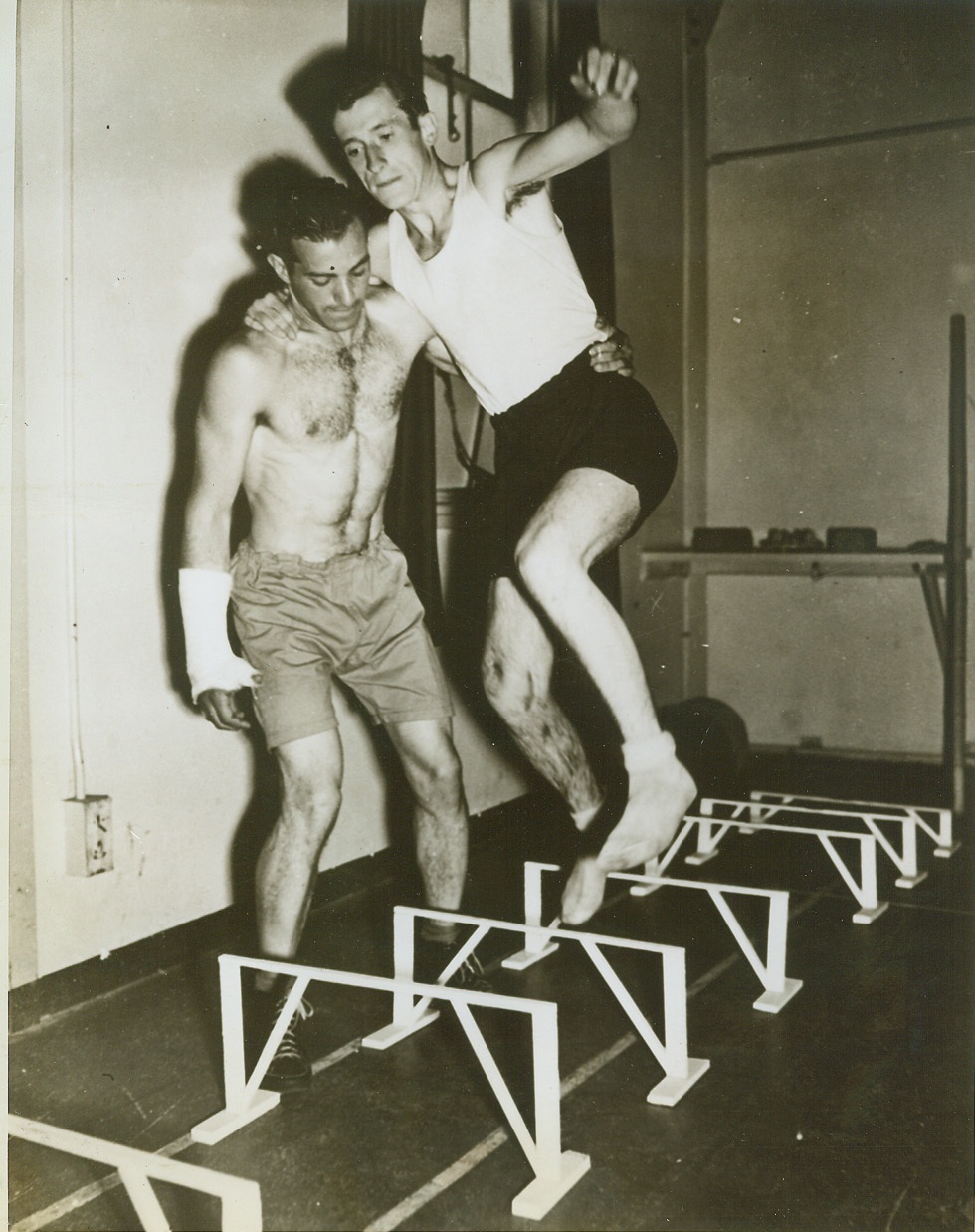 U.S. Soldiers Run Painful Race to Health, 6/26/1943. Getting over these foot high hurdles is no easy task for Pvt. Ralph Johnson, of Pittsburg, Pa., who needs one strong arm of Sgt. D.D. Gilbert of Philadelphia, Pa., to finish out his first race with health. Hospitalized somewhere overseas these wounded American soldiers must take special therapeutic exercises in the hospital gymnasium to rehabilitate the injured limbs and muscles. Credit: (Signal Corps Photo from OWI – ACME);