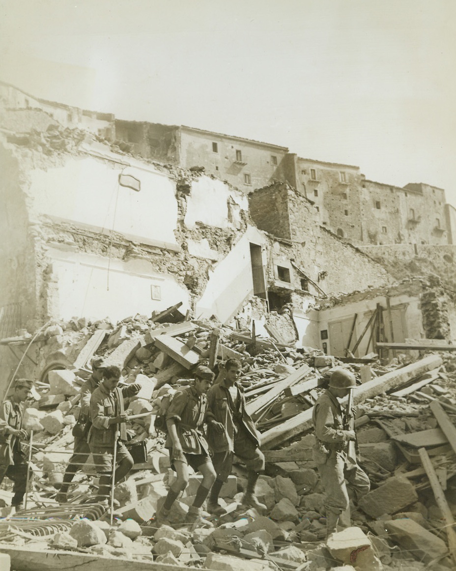 THE WAR IS OVER FOR THEM, 8/14/1943. SICILY—A U.S. soldier leads a group of captive Italian soldiers through the debris that clutters the streets of bomb-scarred Troina after our troops pushed the Axis out of the town. They are on their way to a temporary internment camp. Note the two pictures still hanging on the wall in the background. They are all that remain as evidence that the building was once a home. Credit: Acme;