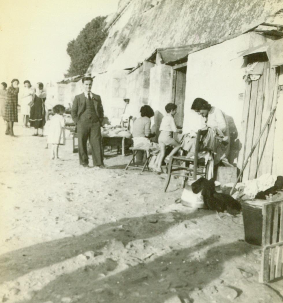 Happy Homes – And Safe, 1/18/1943. Malta – Although there is little left of Malta’s cities after innumerable bombings, but piles of rubble, the Maltese have retained their fine spirit. Here, in front of homes hewn out of the walls of an old fort, people of Malta go happily about their daily tasks in the warm sunshine. Several Maltese in background, seem amused at having their picture taken. 1/18/43 Credit Line (ACME);