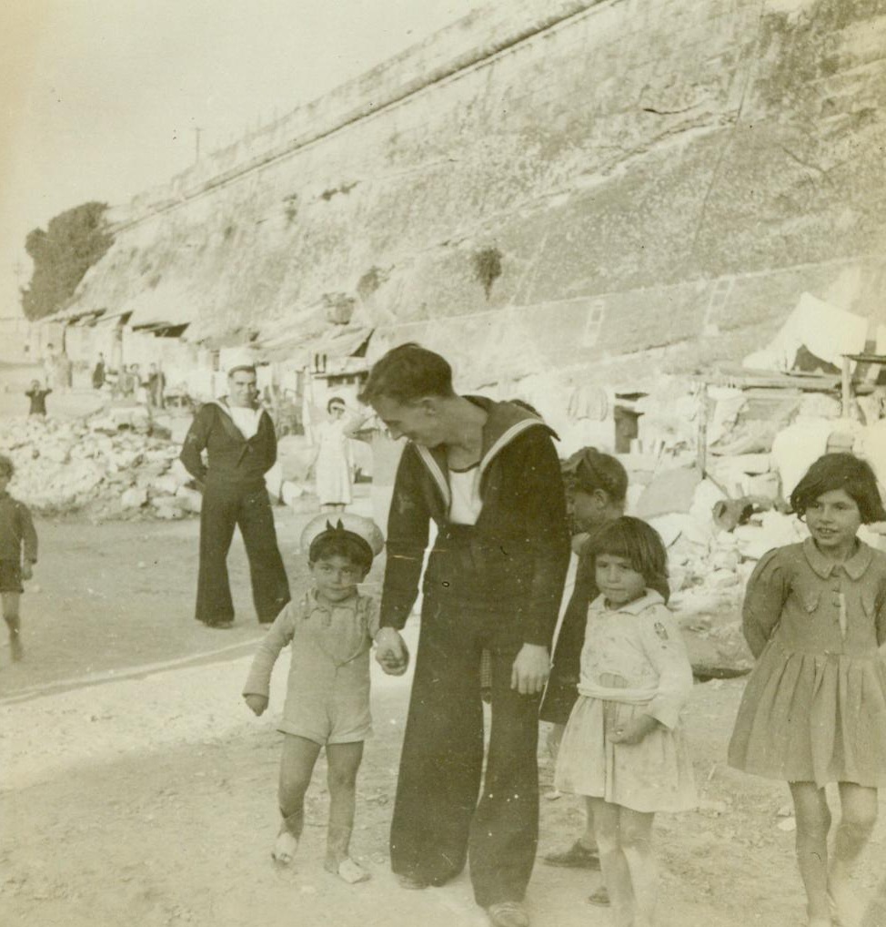 Malta’s Children Can Still Smile, 1/18/1943. Malta – In spite of the fact that the island of Malta has been bombed more than any other place on earth, its people still can smile. Here, Maltese children enjoy a few moments with British Sailors. In background, is an old fort where homes and tunnels have been hewn from the walls for use as shelters during air raids. 1/18/43 Credit Line (ACME);